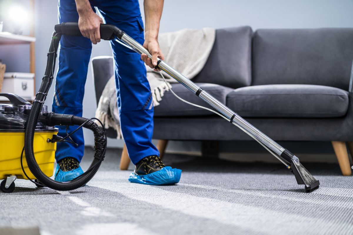 A person in blue scrubs uses a vacuum cleaner on carpeted flooring, wearing protective blue shoe covers, near a grey sofa.