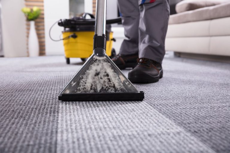A person cleans a carpet using a professional cleaning machine, revealing dirty water in the cleaning head. A yellow cleaning tank is nearby.