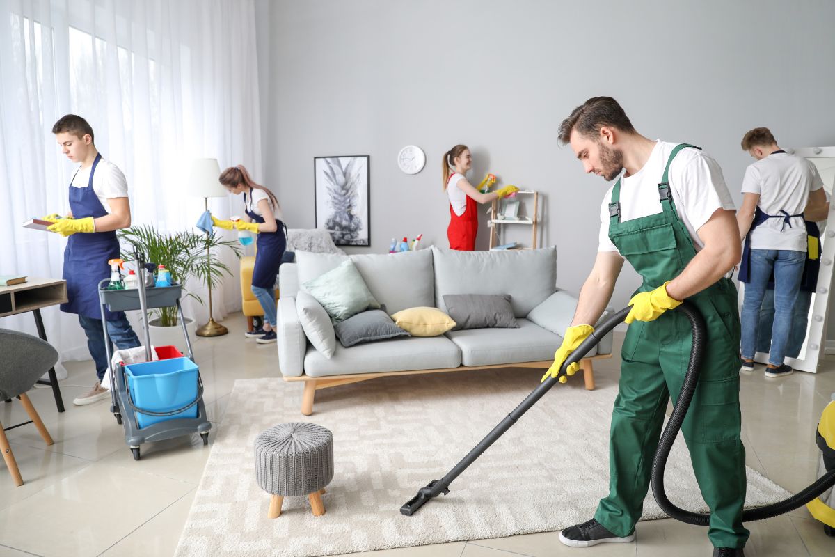 A group of four individuals clean a modern living room, using a vacuum, cloths, and cleaning supplies in a bright, stylish space.