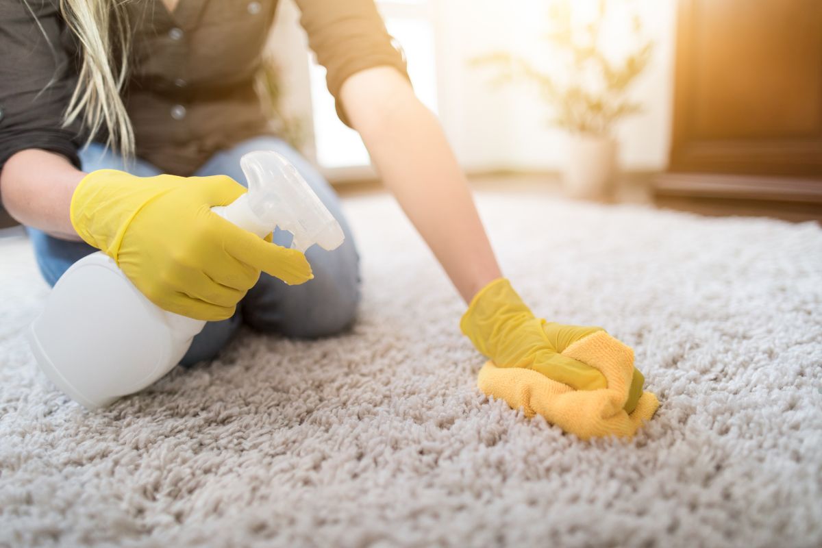 A person in yellow gloves cleans a carpet using a spray bottle and a cloth, focusing on maintaining a tidy home environment.