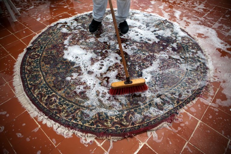 A person sweeps foam and water over a colorful rug on a tiled floor, emphasizing a cleaning process.